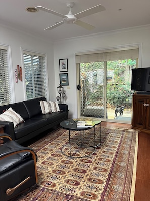 A living room with a black sofa, a coffee table, a patterned rug, a ceiling fan, and a TV on a wooden cabinet. - Light bright and airy, garden outlook from every window, quiet location (Glen Iris)