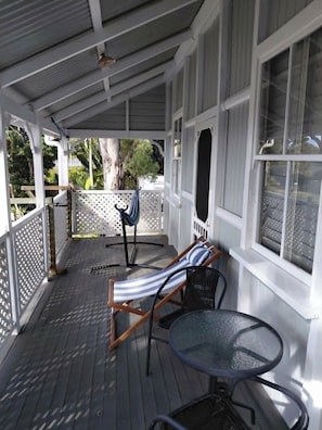 A covered porch with a wooden deck, a lounge chair, a small table, and a view of the outdoors.