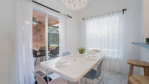 En spiseplads med et hvidt bord dækket til fire personer, stole, en potteplante og et hængende lysarmatur.
