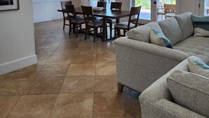 Un vaste espace de vie comprenant un grand canapé gris, une table à manger avec des chaises, un ventilateur de plafond et une vue sur l’extérieur grâce à des portes vitrées.