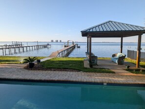Una piscina con un muelle que se adentra en una masa de agua, un pabellón con asientos y una zona verde con plantas en macetas.