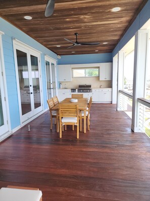 A covered patio area with a wooden table and chairs, a ceiling fan, and a kitchen area with a window. - Your Bayfront Retreat with Stunning Water Views, Private Pool, and Dock. (Gulf Breeze)