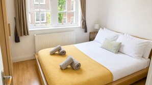 A hotel room with a bed, a window with curtains, a radiator, and a nightstand with a lamp. - 2bed Home With Free Parking Near Sydenham Station (London)