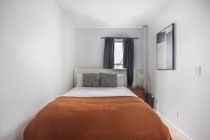 Ein schmales Hotelzimmer mit Bett, Nachttisch, einem Fenster mit Vorhängen, einer Heizung und einem gerahmten Bild an der Wand.