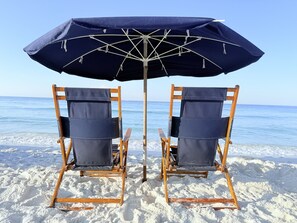 Two wooden beach chairs with canvas seats and backs, positioned under a blue umbrella on a sandy beach with the ocean in the background. - Beach Front Paradise/Unobstructed Gulf View From Private Balcony/Pool (Destin)