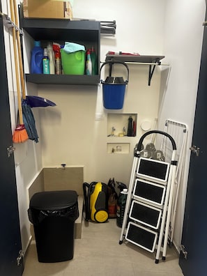 A small, well-organized utility room with shelves holding cleaning supplies, a mop, a broom, a trash bin, and a ladder.