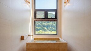 A bathroom with a bathtub, a window with a view of a forest, and wall-mounted lighting.