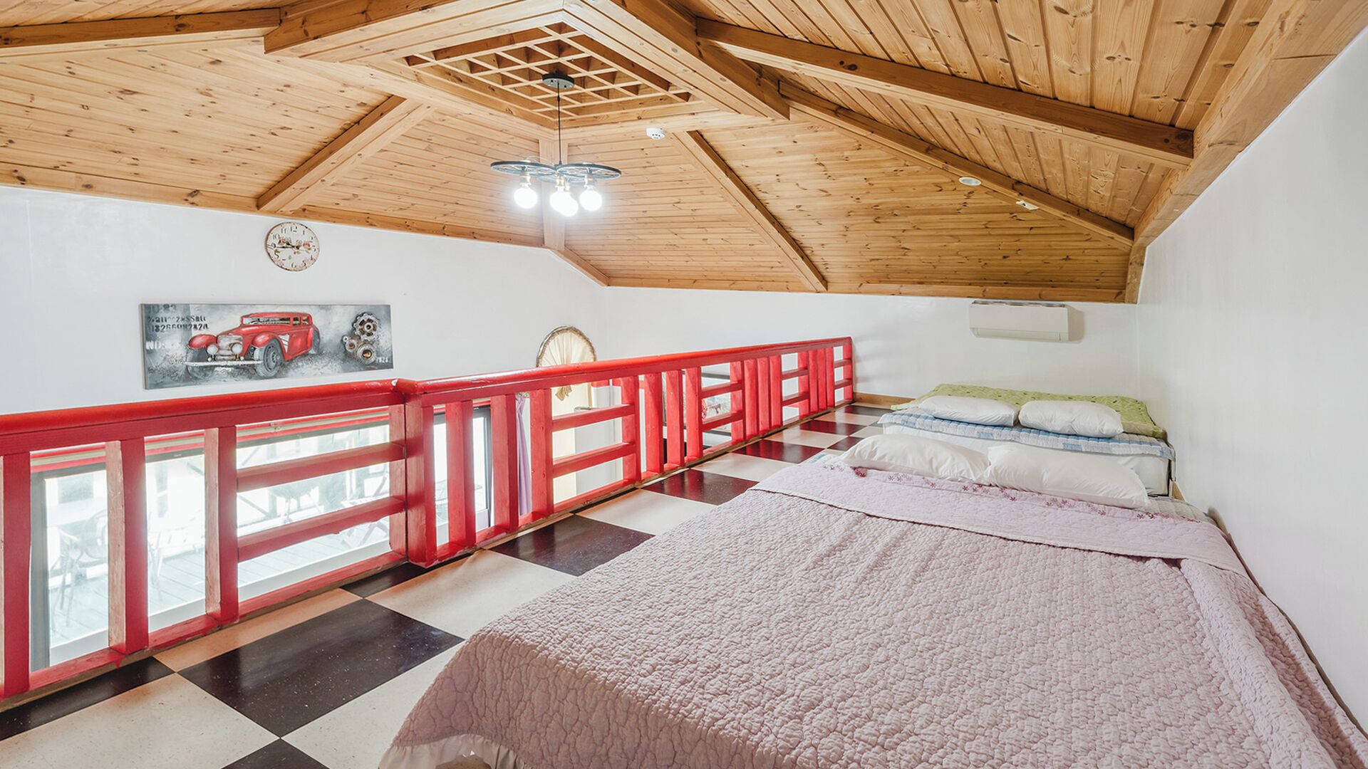 A bedroom with a checkered floor, a bed with a pink blanket, a red railing, a wooden ceiling, and a clock on the wall.