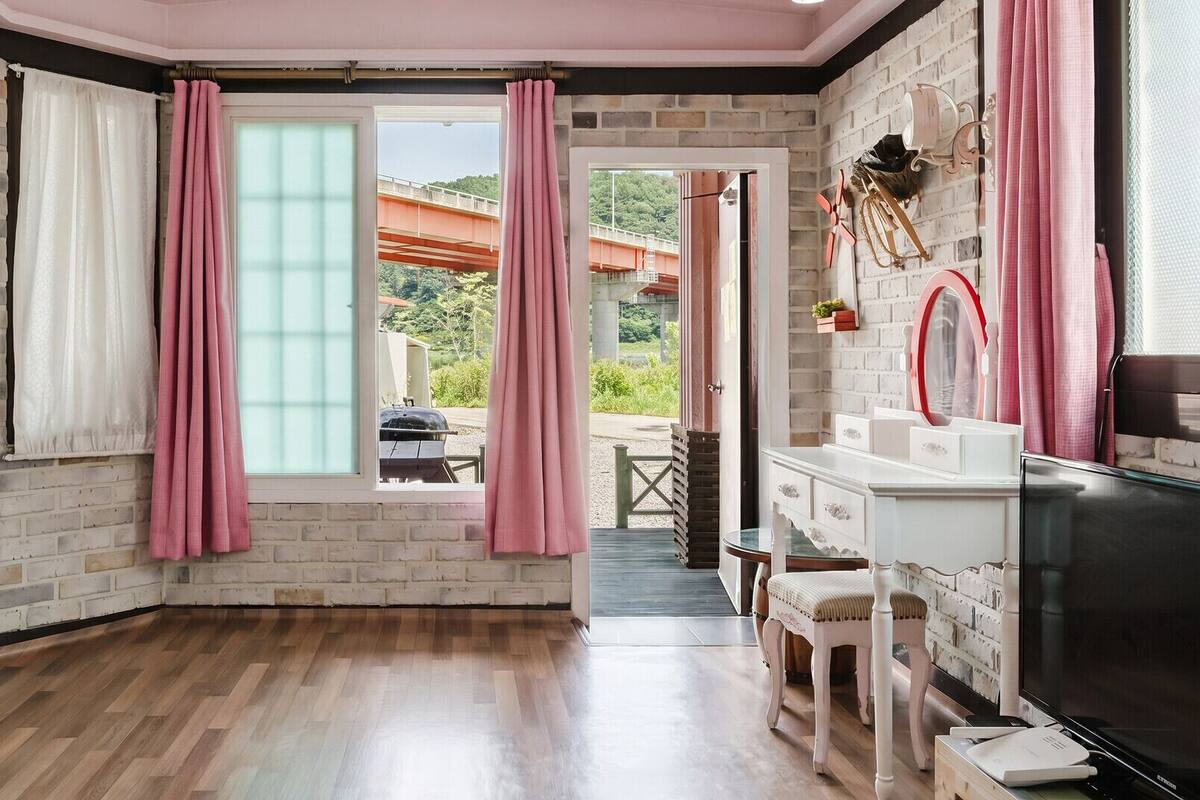 Una habitación con pared de ladrillo, suelo de madera, televisor, tocador con espejo y cortinas rosas.