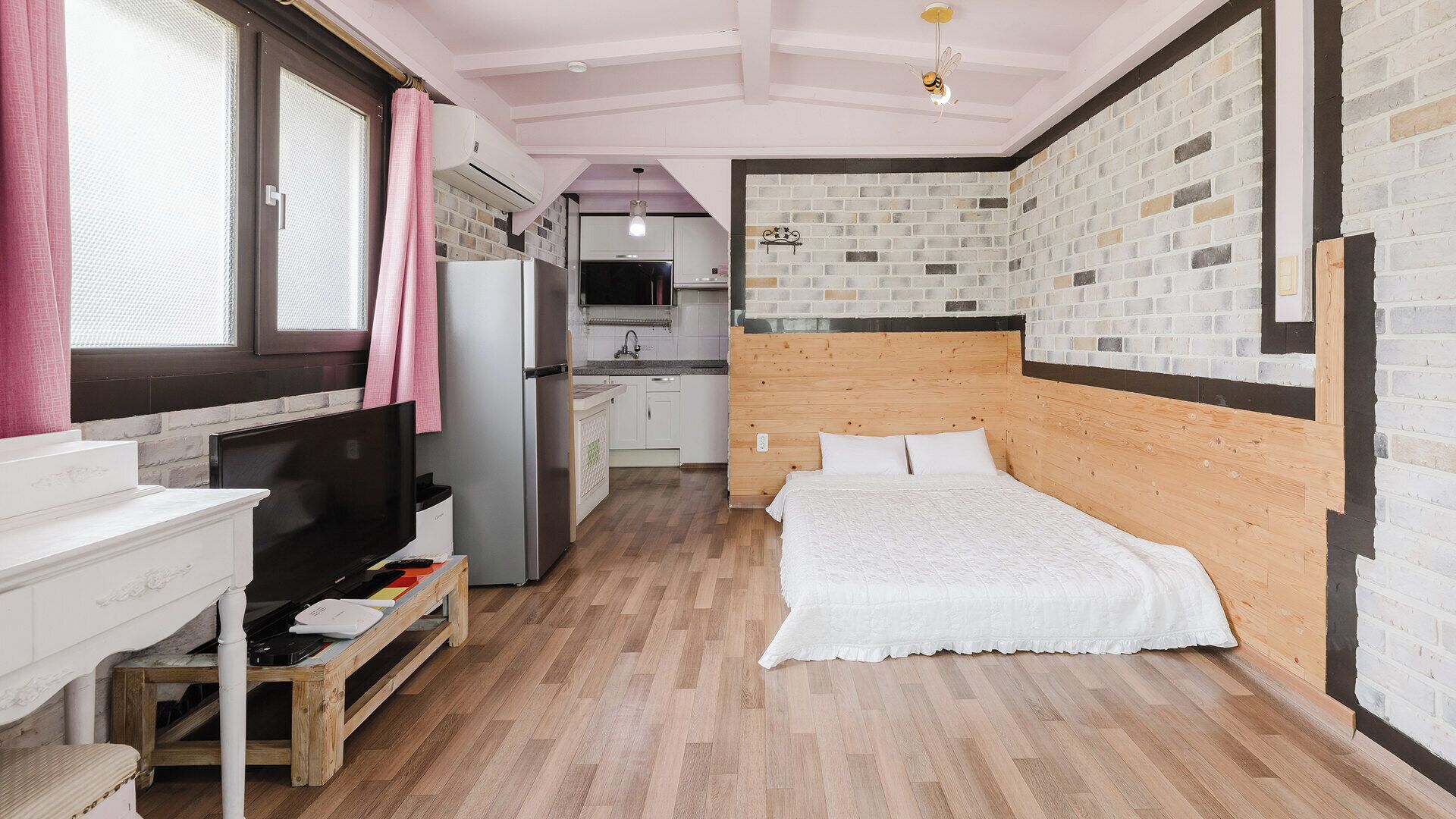 A compact bedroom with a wooden headboard, a bed, a desk with a chair, a television, and a kitchen area with a refrigerator and sink.