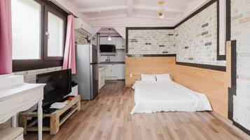 A compact bedroom with a wooden headboard, a bed, a desk with a chair, a television, and a kitchen area with a refrigerator and sink.