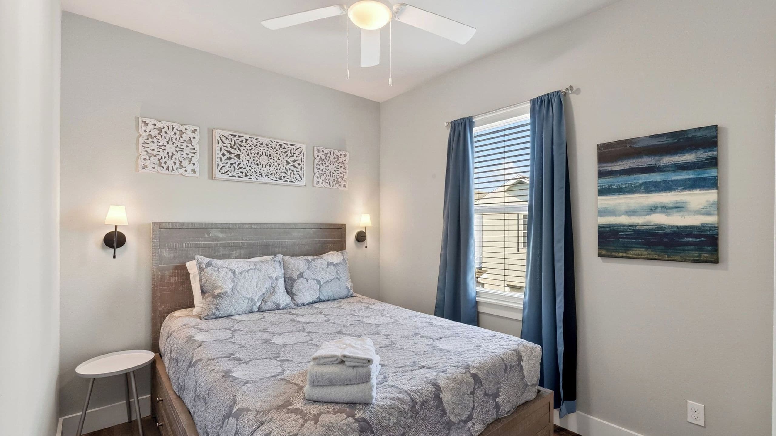 A bedroom with a bed, a ceiling fan, decorative wall art, a window with curtains, and a painting on the wall.