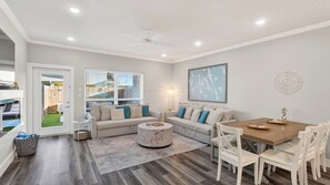 Ein modernes Wohnzimmer mit einer Sofagarnitur, einem runden Couchtisch, einem Esstisch mit Stühlen, einem Deckenventilator und einem Fenster mit Jalousien.