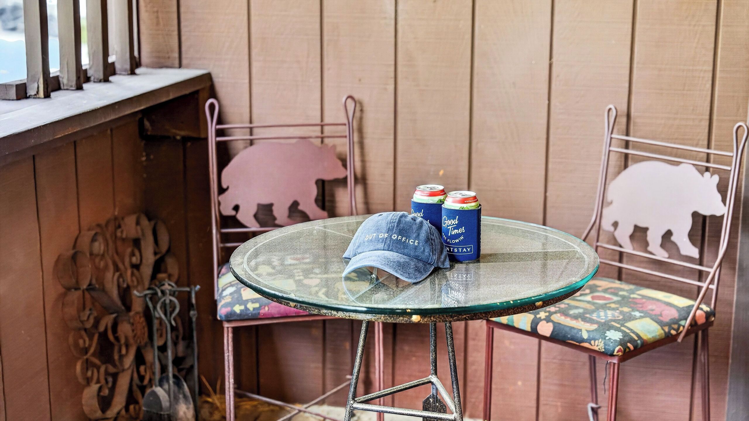 Una mesa de vidrio con dos latas y una tapa, sillas de metal con recortes de animales, y una pared de madera con una silueta de cerdo.