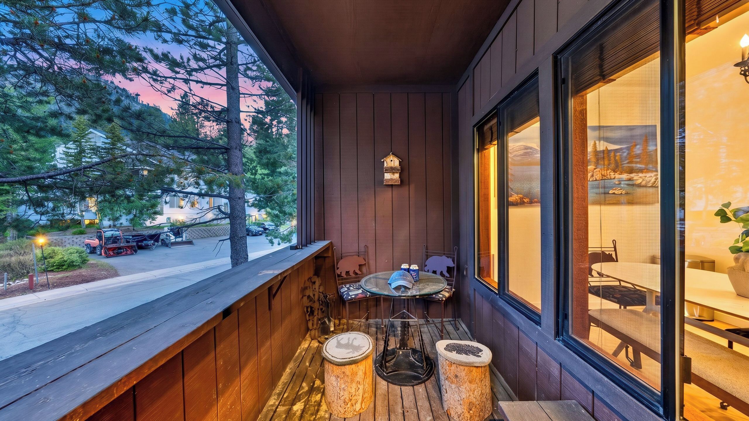 A wooden balcony with a table and chairs, a birdhouse, and a view of a road and trees.