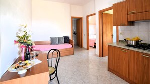 A compact room with a bed, a dining table with a chair, a kitchenette, and a bathroom visible through an open door. - Apartment in Ricadi (049) (Ricadi)