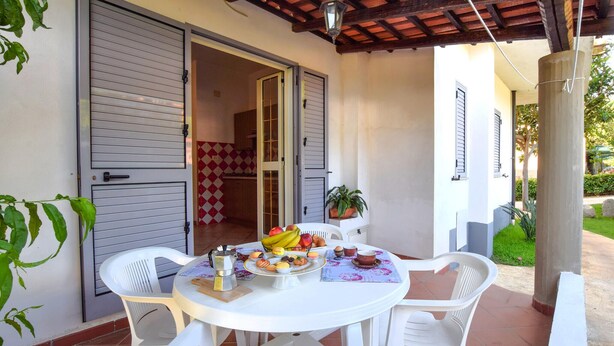 Een terras met een ronde tafel gedekt voor het ontbijt, witte plastic stoelen en uitzicht op het interieur van het huis.