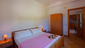 Een slaapkamer met een bed, nachtlampjes, een houten kledingkast en een kitchenette zichtbaar door een geopende deur.