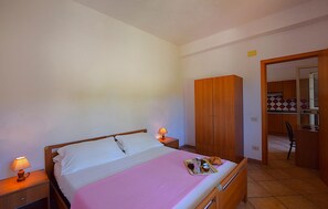 Une chambre à coucher avec un lit, des lampes de chevet, une armoire en bois et une kitchenette visible à travers une porte ouverte.