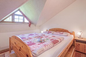 Quarto com teto inclinado, cama de madeira, mesa de cabeceira com abajur e janela com vista para árvores.