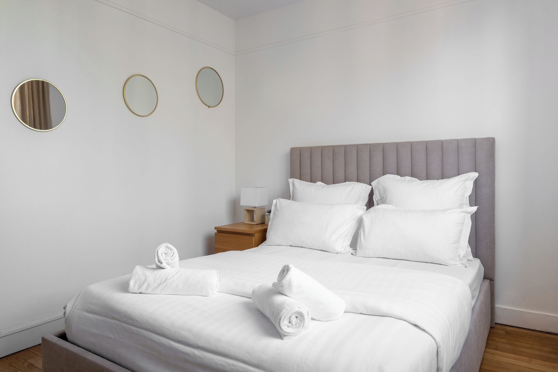 Uma cama bem arrumada com lençóis e almofadas brancos, uma cabeceira estofada cinza e uma mesa de cabeceira de madeira com abajur.