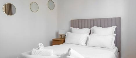 Una cama bien hecha con sábanas y almohadas blancas, un cabecero tapizado de gris y una mesita de noche de madera con una lámpara.