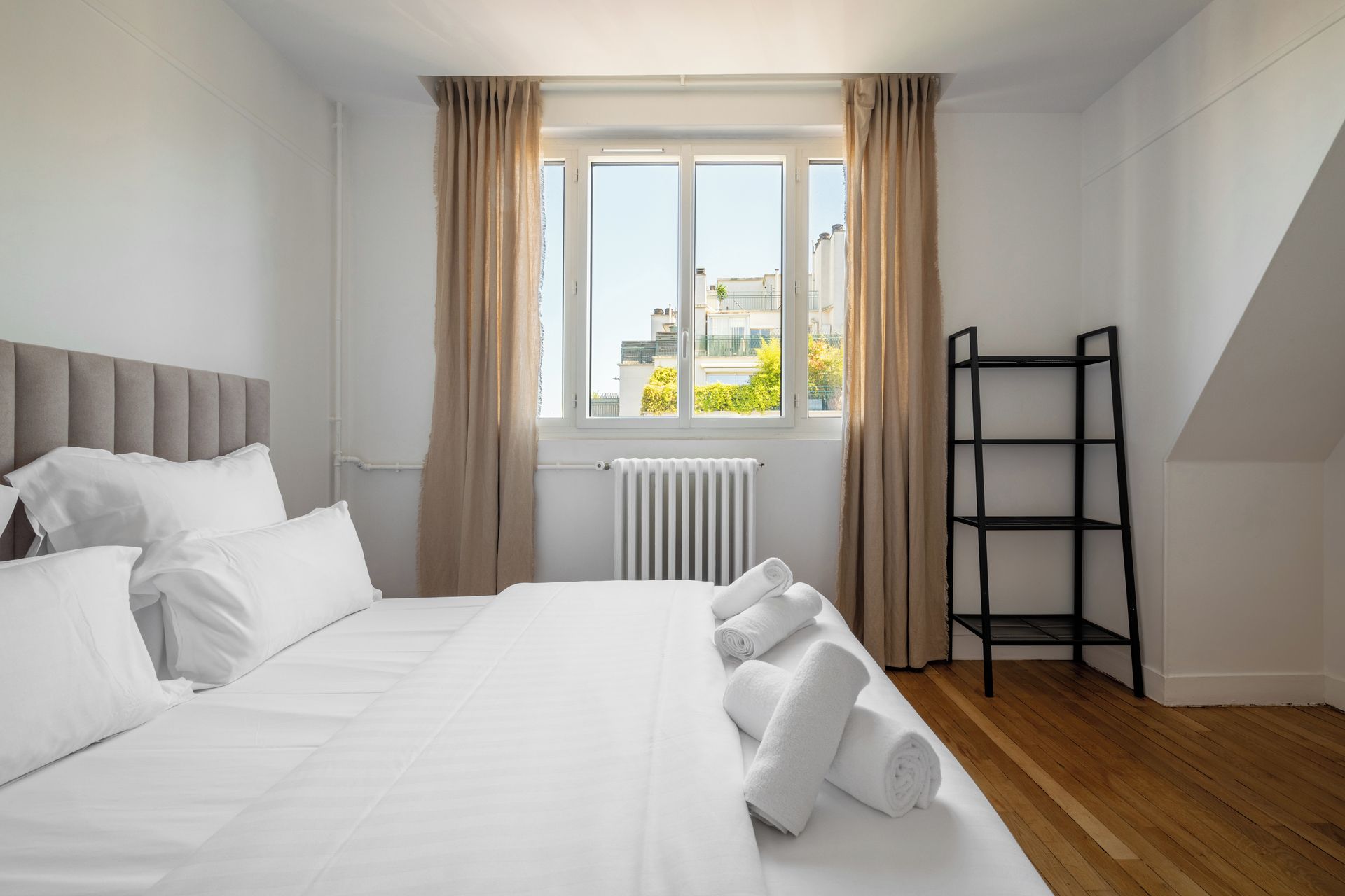 A hotel room with a bed, a radiator, a window with curtains, and a wooden floor.