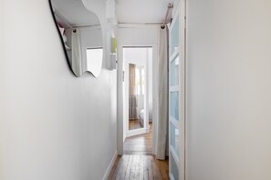 A narrow hallway with a wooden floor, white walls, a mirror, and a door leading to a room with a bed. - Bright 1BDR Retreat in Paris 15th (Paris)