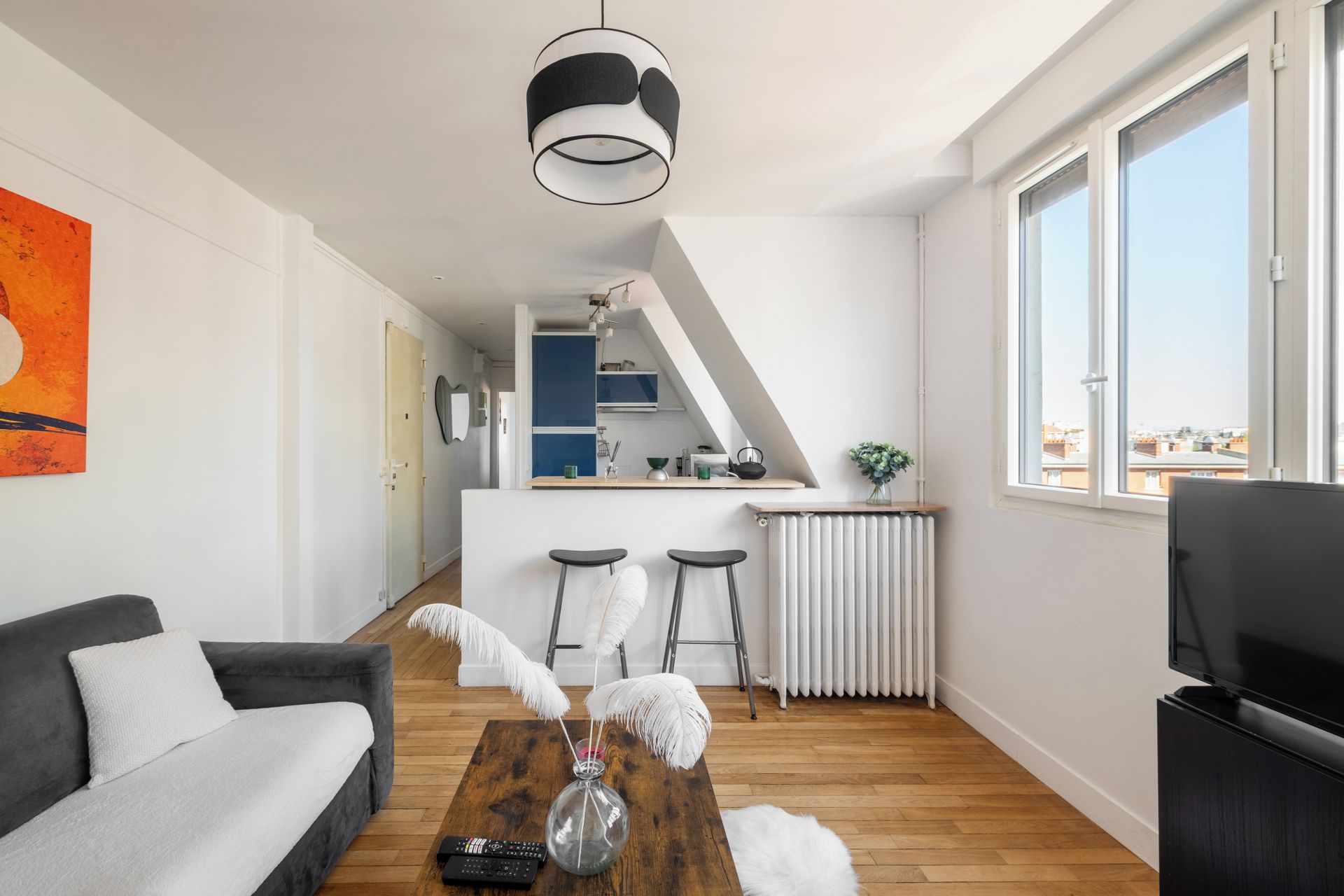 Un salon moderne avec un canapé gris, une table basse en bois et un téléviseur. On aperçoit, au fond, un coin cuisine avec une fenêtre donnant sur la ville.