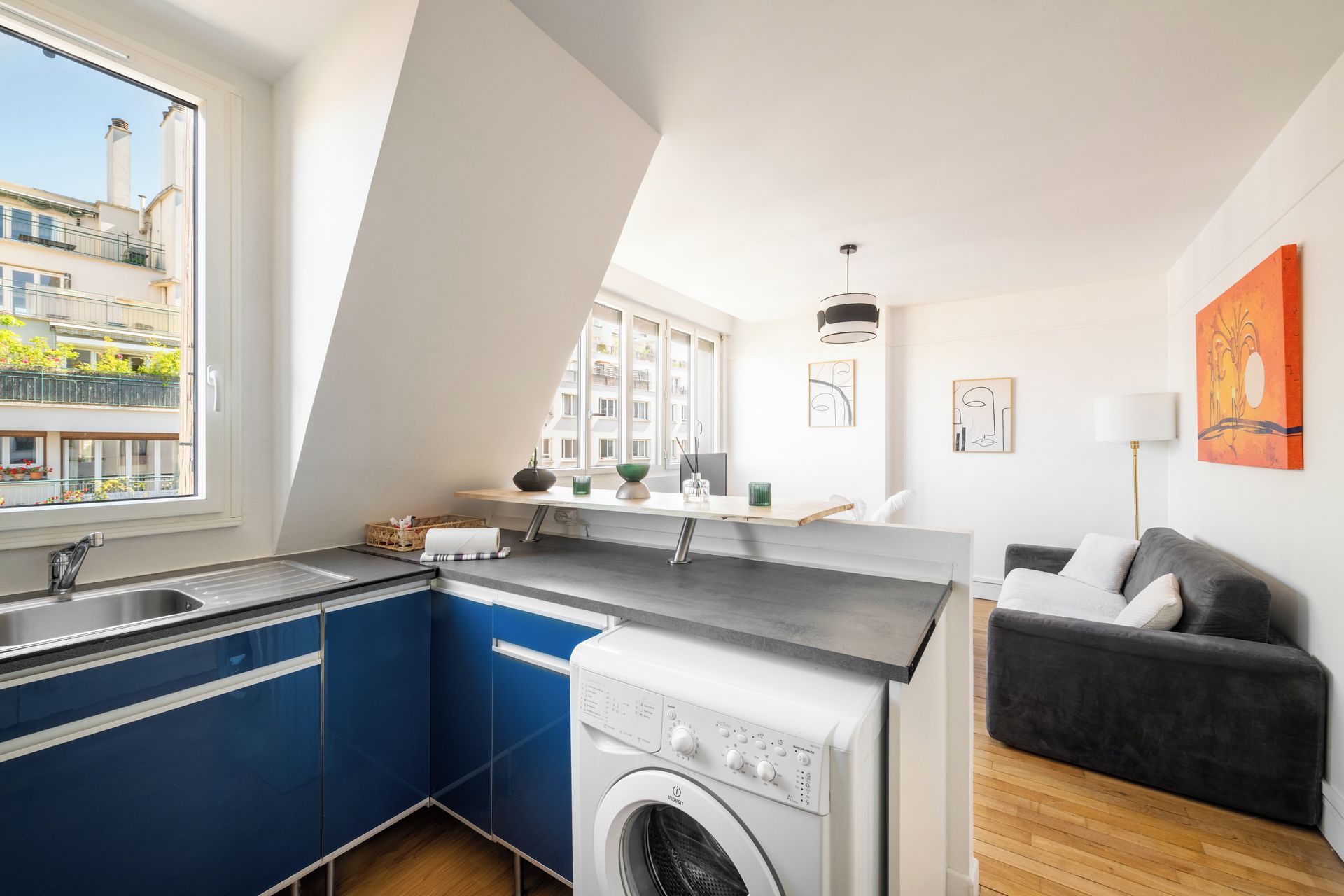 A compact kitchen with a built-in washing machine, a window with a view of a balcony, and a grey sofa in the living area.