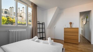 Een slaapkamer met een bed, een radiator, een raam met gordijnen, een houten dressoir en een ladder.