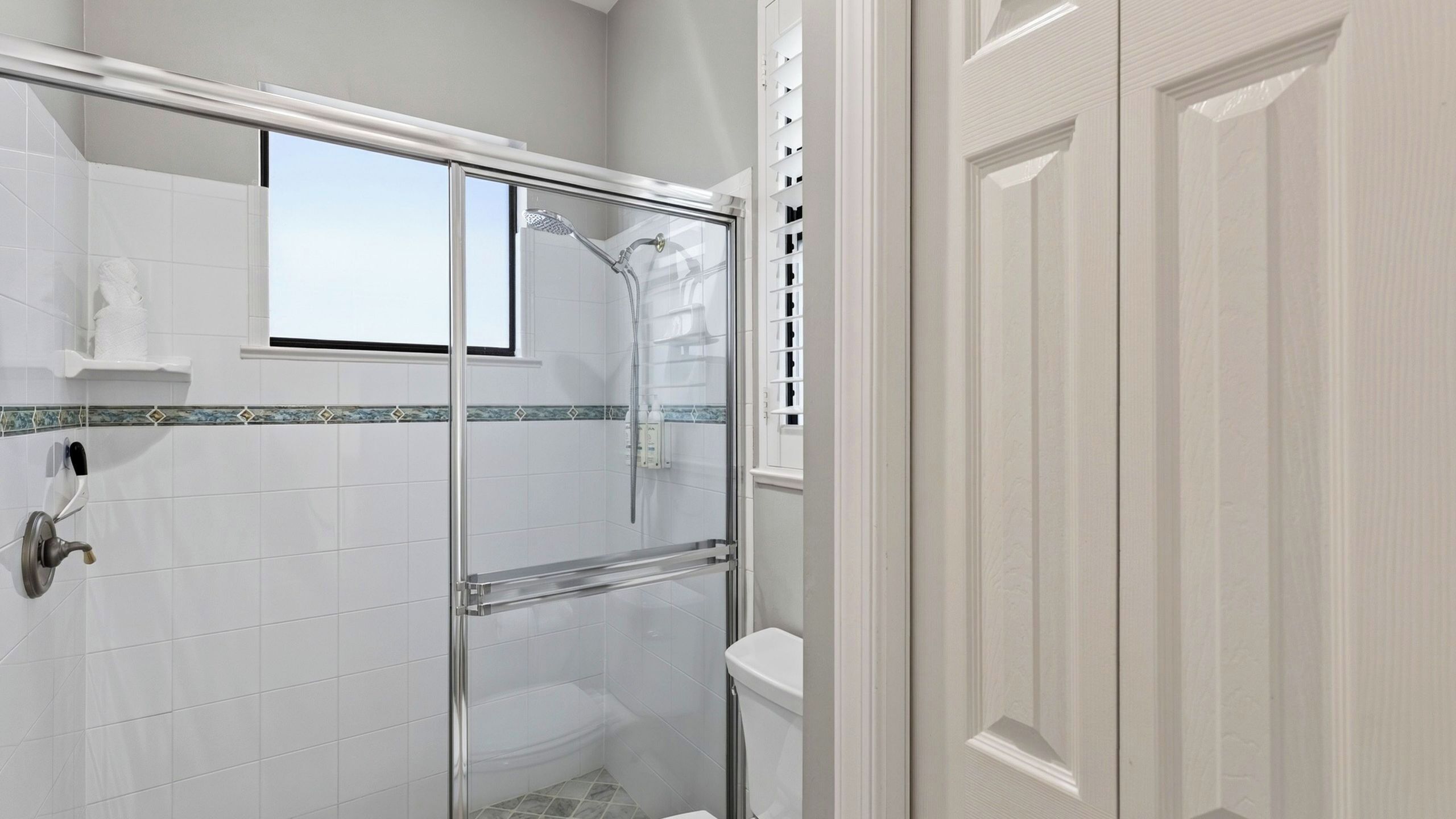 Ein Badezimmer mit Duschkabine aus Glas, weiß gefliesten Wänden, Toilette und Fenster.