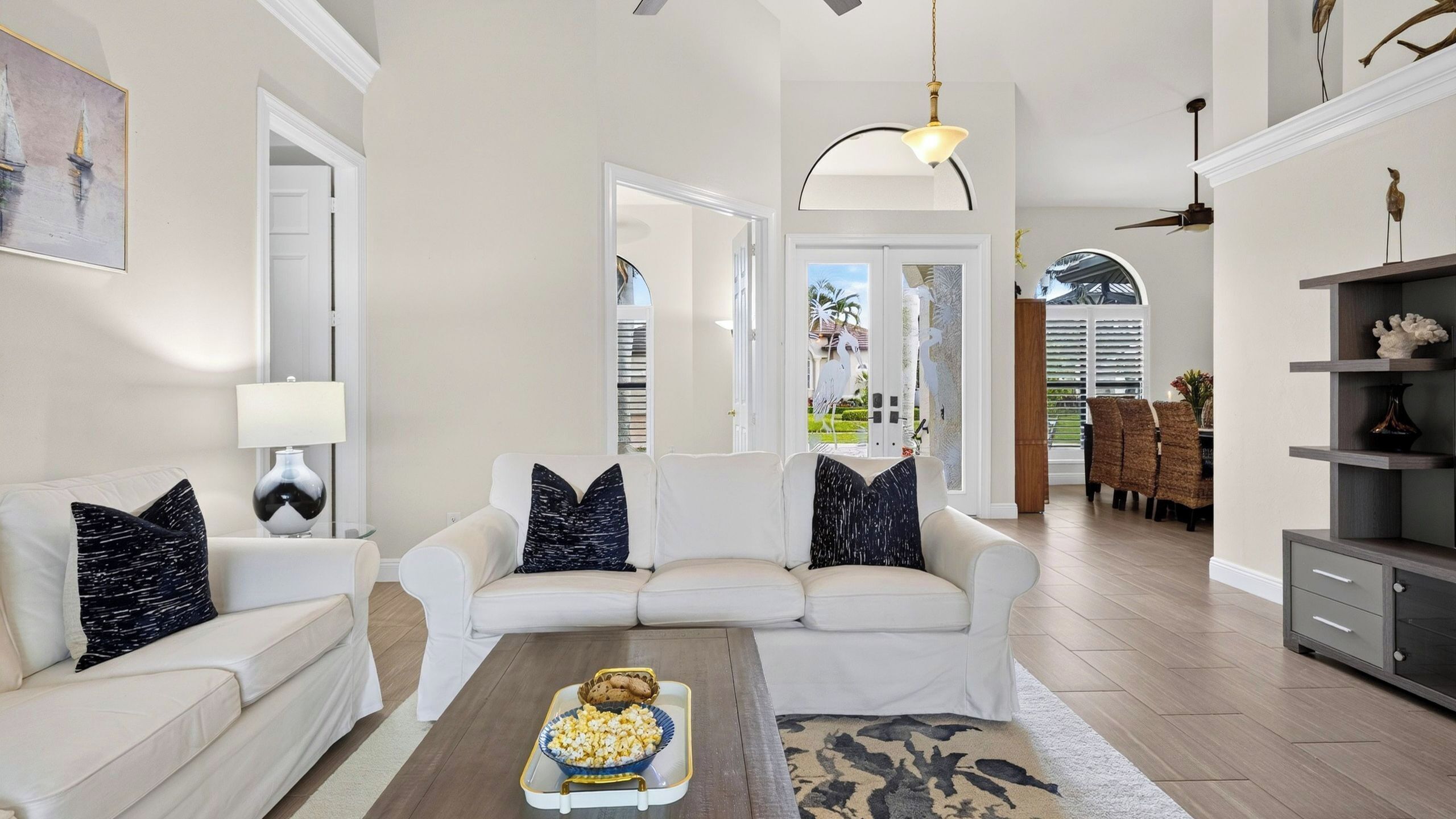 A spacious living room with a white sofa set, a coffee table with snacks, and a view of the outdoor patio.
