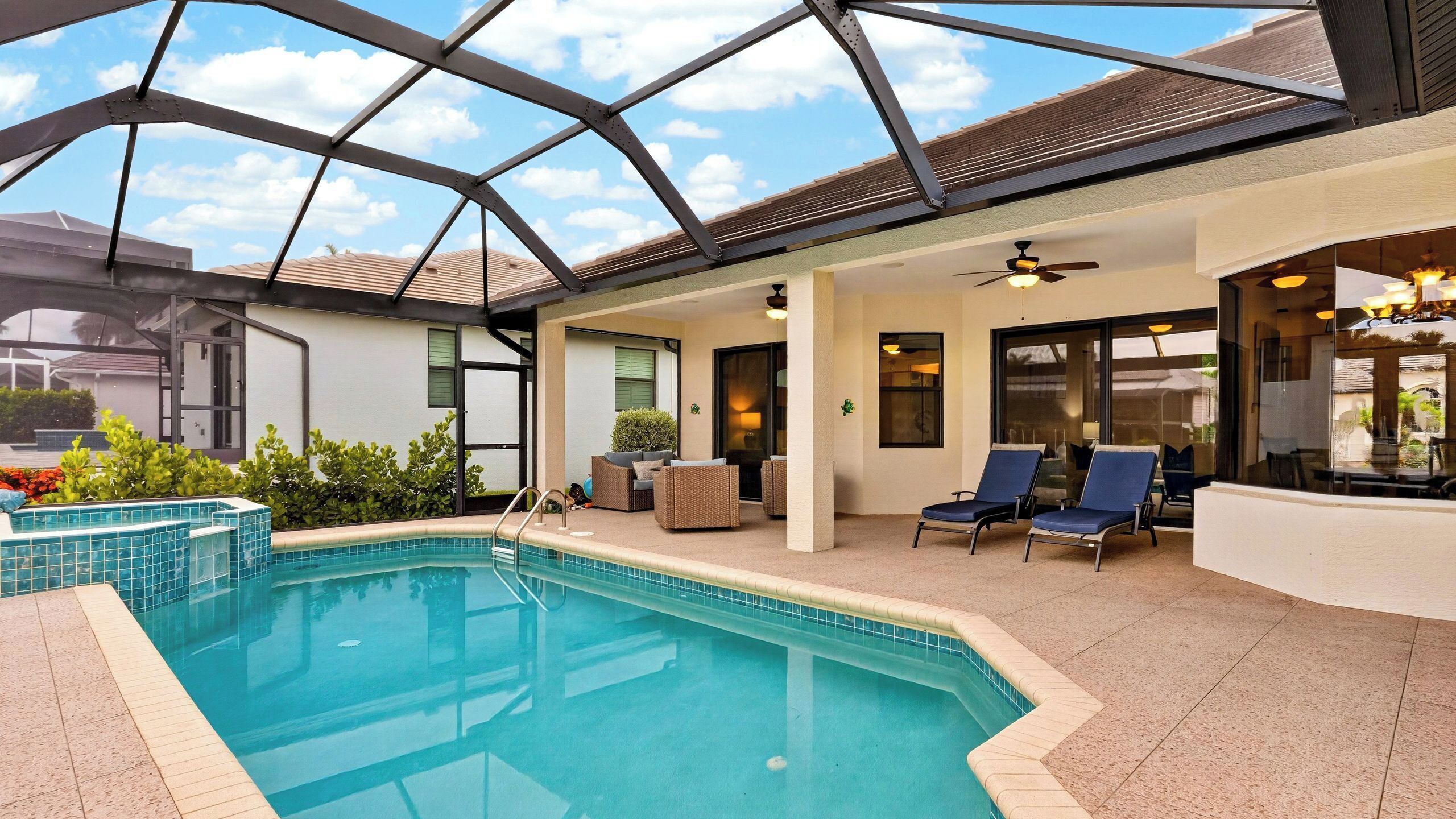 A covered outdoor swimming pool with lounge chairs, a ceiling fan, and a glass door leading to a patio area.