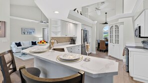 A modern kitchen with white cabinetry, a marble island, and ceiling fans. - Neptune's Realm by Avantstay Waterfront Home + Heated Pool + Spa + Dock Minutes to Beach (Marco Island)