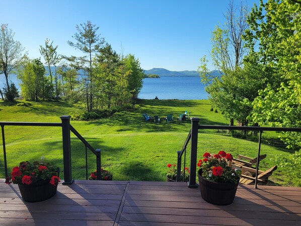 A deck with a view of a lake, surrounded by greenery and a distant windmill. - Island View Escape: Lake, Private Beach, Deck, Firepit, Family, Game Room, more! (Broadalbin)