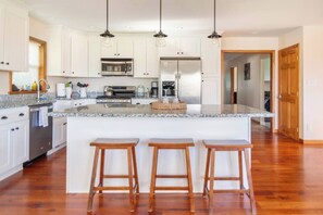 A kitchen with white cabinets, a large island with stools, stainless steel appliances, and pendant lighting. - Island View Escape: Lake, Private Beach, Deck, Firepit, Family, Game Room, more! (Broadalbin)