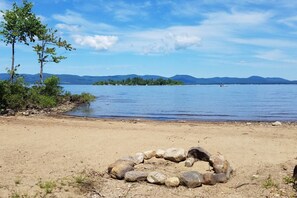 A lakeside scene with a clear sky, a sandy beach, and a ring of rocks in the foreground. - Island View Escape: Lake, Private Beach, Deck, Firepit, Family, Game Room, more! (Broadalbin)