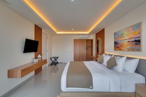 A modern hotel room with a flat-screen TV, a bed with white bedding, a wooden bench, and a painting on the wall. - The Queeno Hill Villa - Nusa Dua (Nusa Dua)