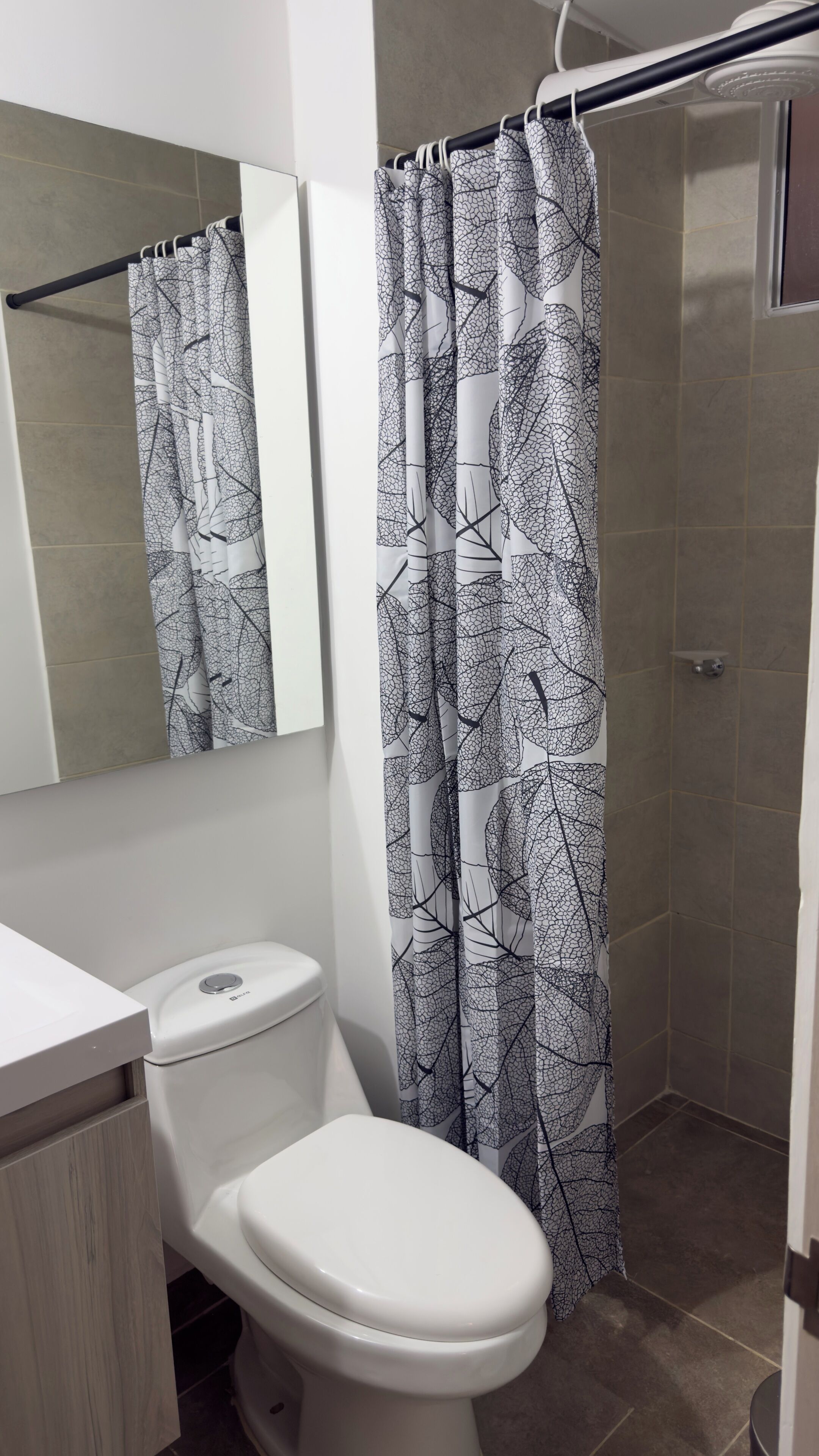 A bathroom with a toilet, a shower curtain with a leaf pattern, a mirror, and tiled walls.