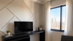 A modern living room with a flat-screen TV, a dark sofa, a small plant, and a city view through a window. - Premium apartment in Tatuapé with pool, gym and garage included. (São paulo)