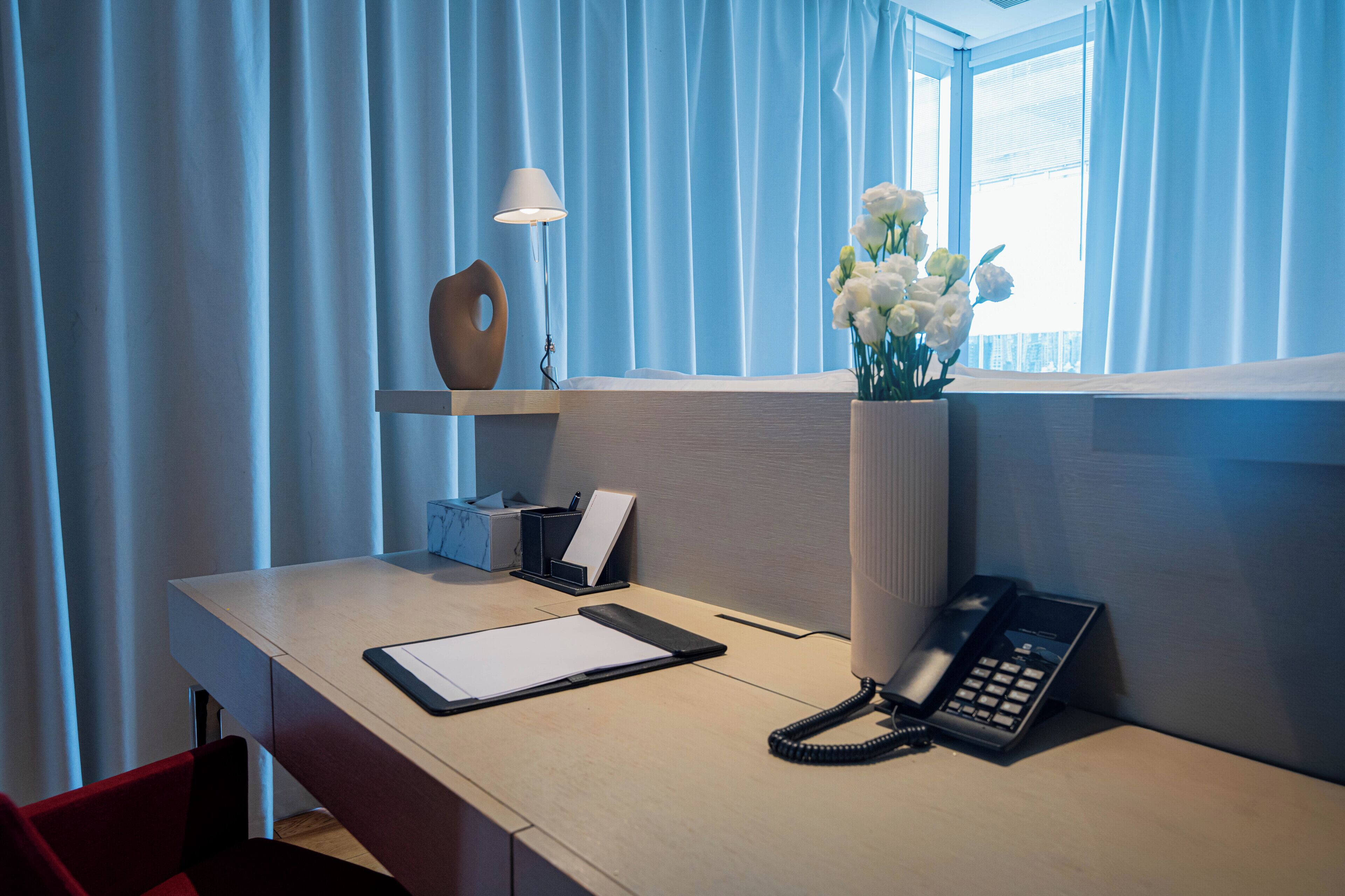 Quarto de hotel com uma escrivaninha, telefone, luminária, um vaso com flores e uma janela com cortinas.