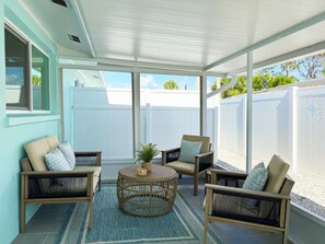 A covered patio with wicker furniture, a central round table, and a view of a white fence and greenery.