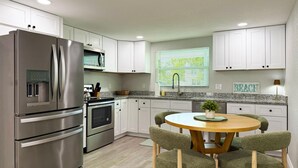 A modern kitchen with stainless steel appliances, white cabinetry, a granite countertop, and a round wooden table with green upholstered chairs.