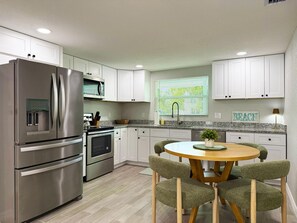 A modern kitchen with stainless steel appliances, white cabinetry, a granite countertop, and a round wooden table with green upholstered chairs.