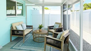 Ein überdachter Patio-Bereich mit Korbmöbeln, einem gewebten Couchtisch und Blick auf einen weißen Zaun und Grünflächen.