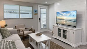 Ein Wohnzimmer mit einer Couch, einem Sessel, einem Couchtisch, einem Fernseher auf einem Schrank und einer Tür mit Jalousien.