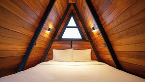 Habitación triangular de madera con una cama, techo inclinado y dos luces fijadas a la pared.