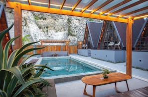 A modern outdoor pool area with a wooden deck, a seating area with chairs, and a potted plant. - EL Cabana (Kuta Selatan)