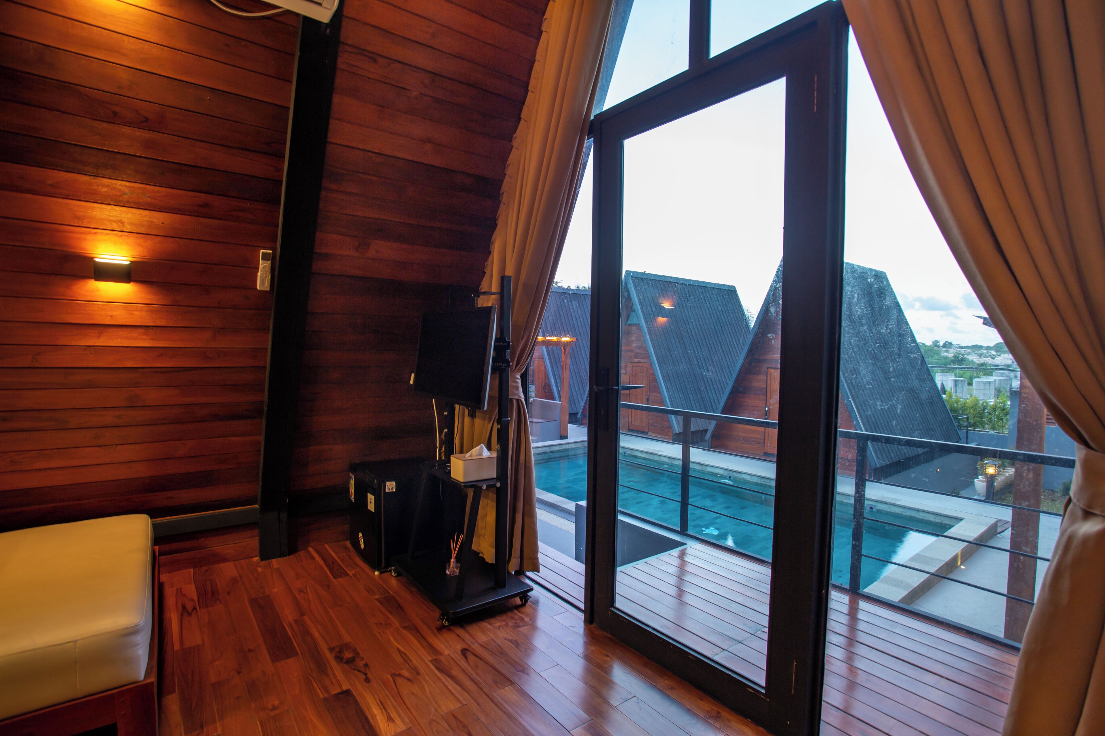 Habitación con paredes de madera, puerta corredera de vidrio a un balcón, un banco blanco y una pequeña mesa con lámpara.